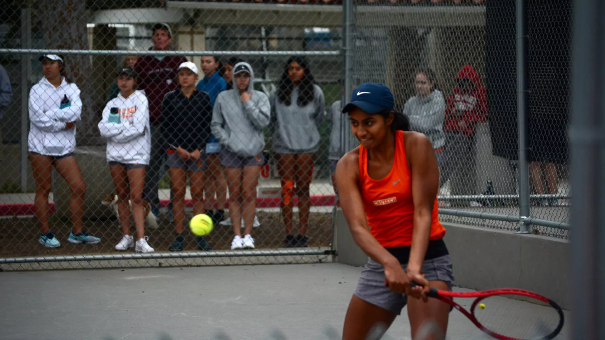 Anika Arora plays her backhand while her teammates support her outside the court