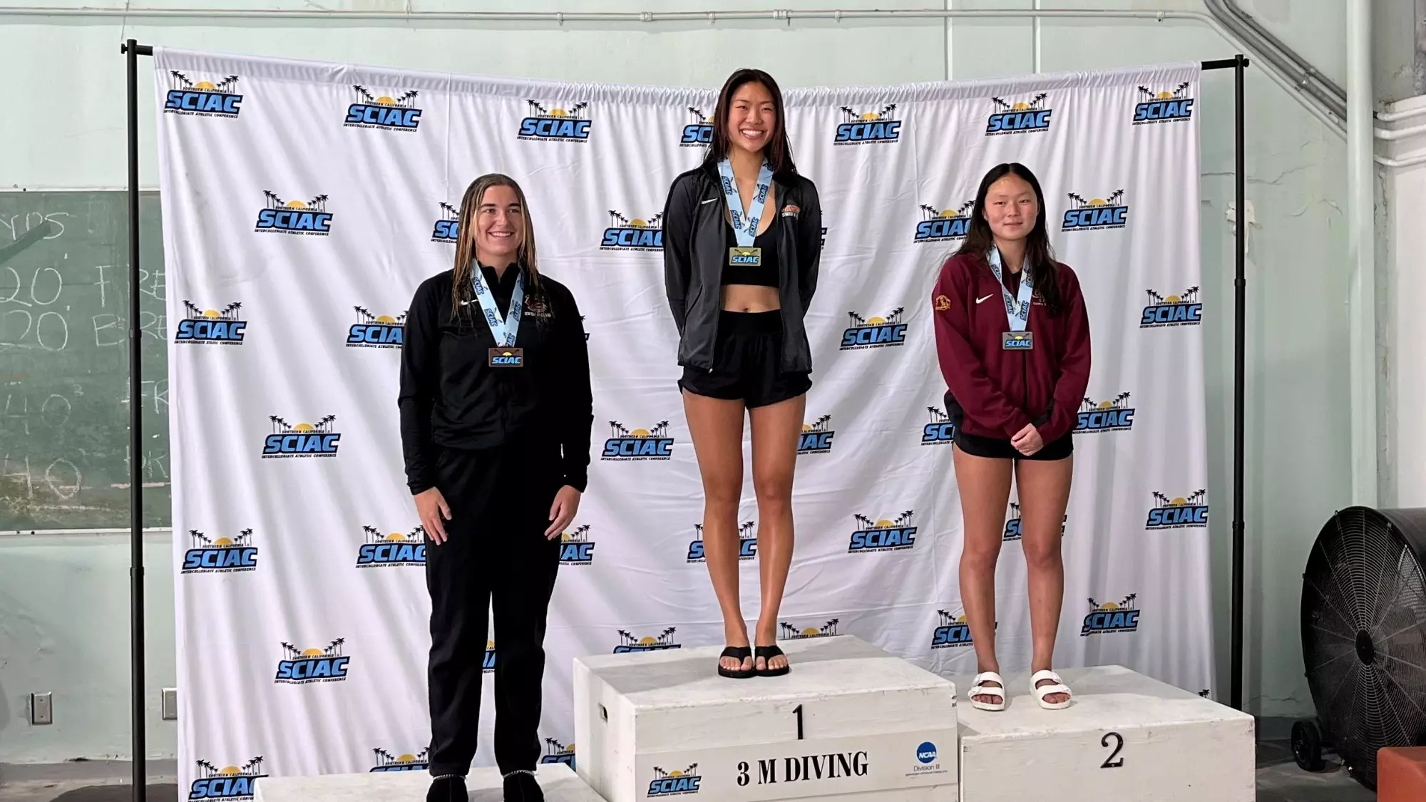 Jana Woo stands on the first place podium between the third place diver from Chapman (left) and the second place diver from Claremont-Mudd-Scripps (right)
