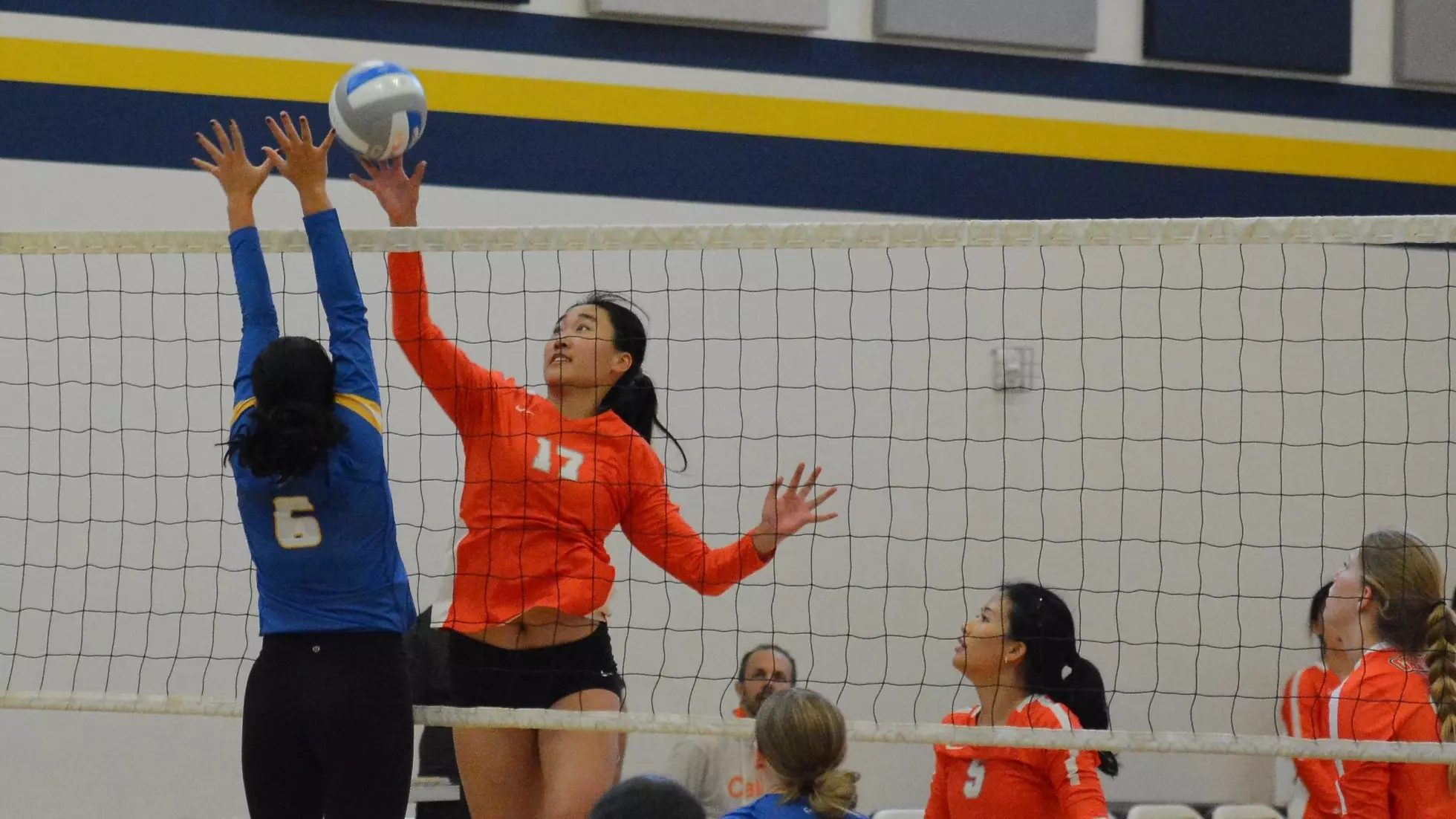 VB 2024 Andrea Li blocking ball in the air above net vs Life Pacific with other players watching in the foreground and background.