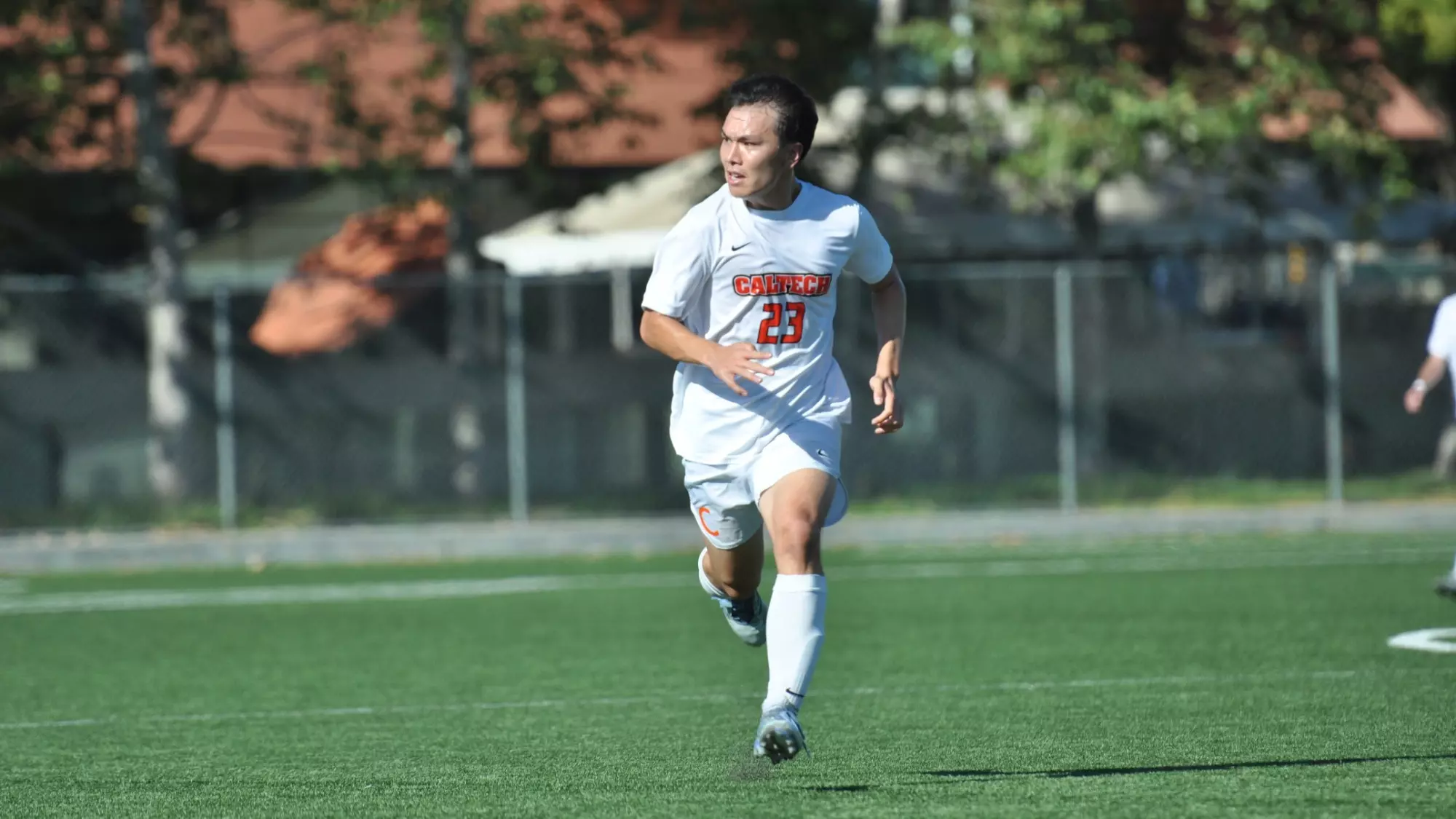 Ethan Feng runs in a white jersey with orange numbers towards the camera. He looks towards his right, the left of the frame, at something out of frame. Both of his feet are off the ground as he is in the middle of his stride. 