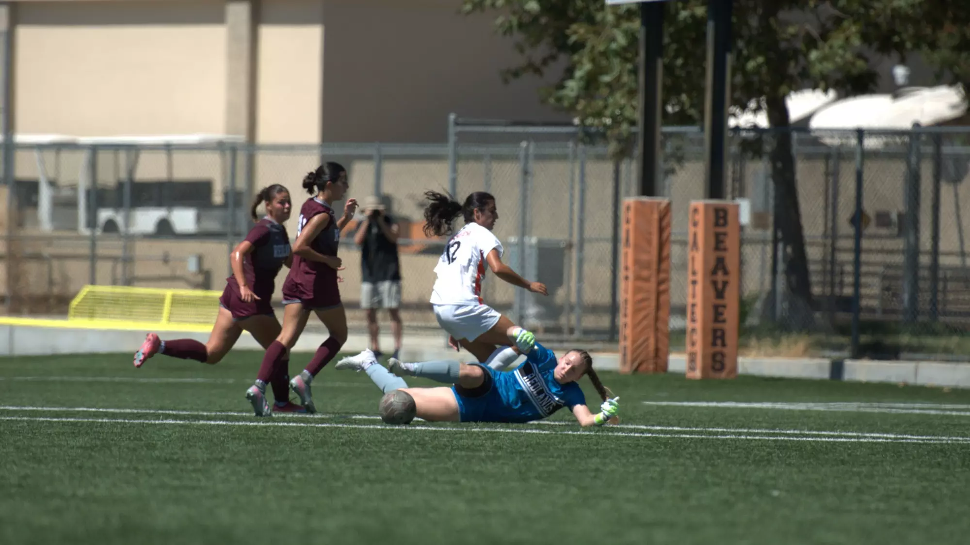 Shyna Kashi, wearing white, strains to stop her momentum after jumping over the goaltender who is sliding on the ground reaching out in an attempt to stop a shot by kashi. Two Redlands defenders, wearing red, attempt to navigate the cluster of bodies. 
