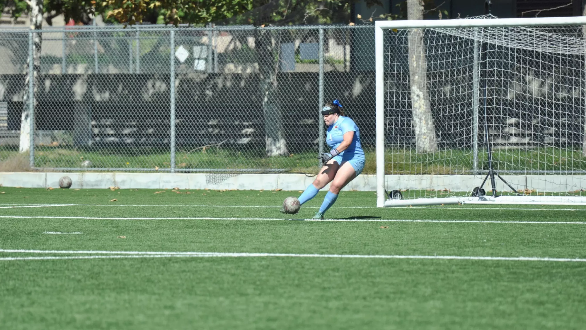 Sophia Steven, wearing an all powder-blue goalie kit, makes contact with the ball while executing a goal kick in front of the right post of the net. The ball has a faded red and grey coloration similar to BB8 from 