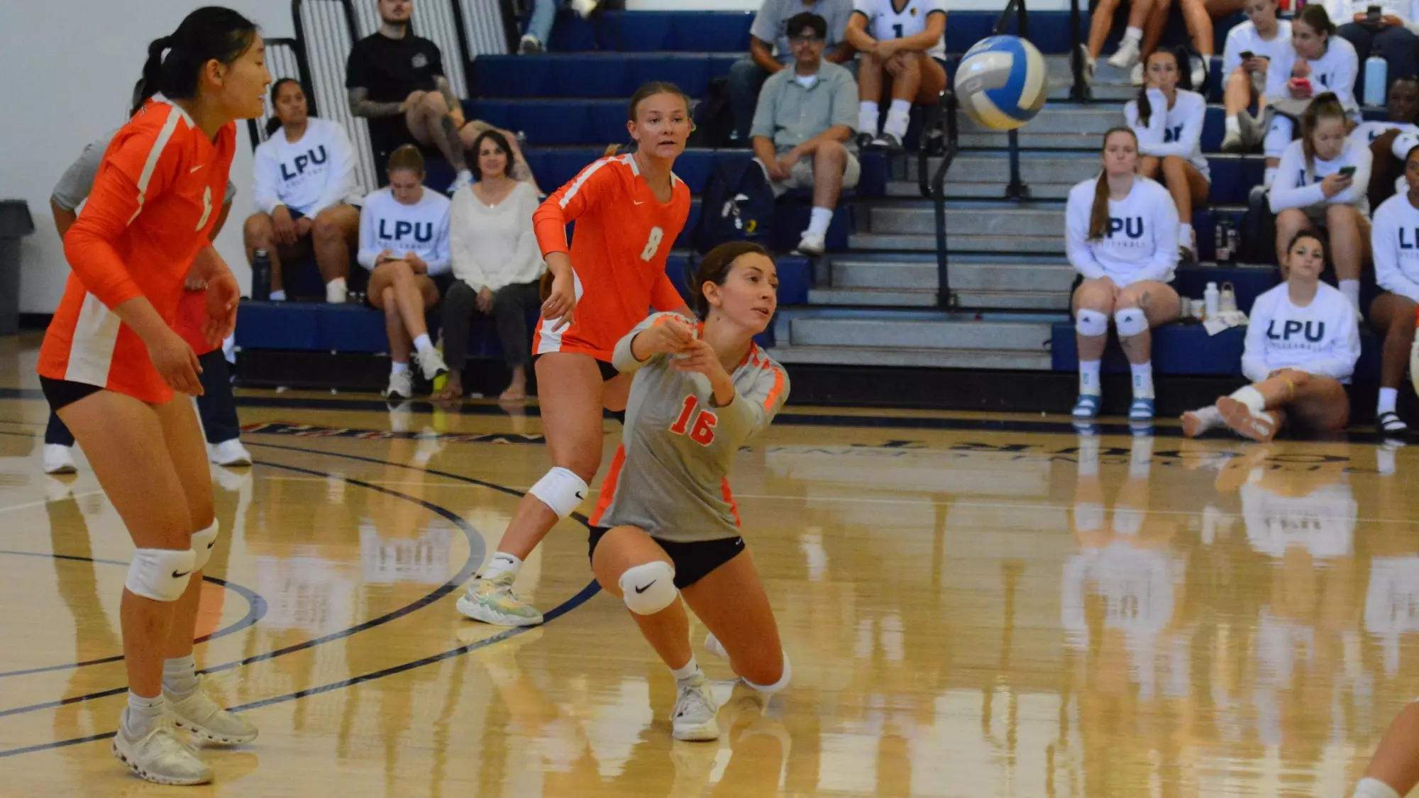 Giulia Murgia lunges to her right with her hands together as she makes a dig against life pacific. She wears an grey libero jersey with orange trim. In orange jerseys with white trim, Rena Zheng and Alize Bakker look on from Murgia's right and left respectively in front of a background of fans wearing 
