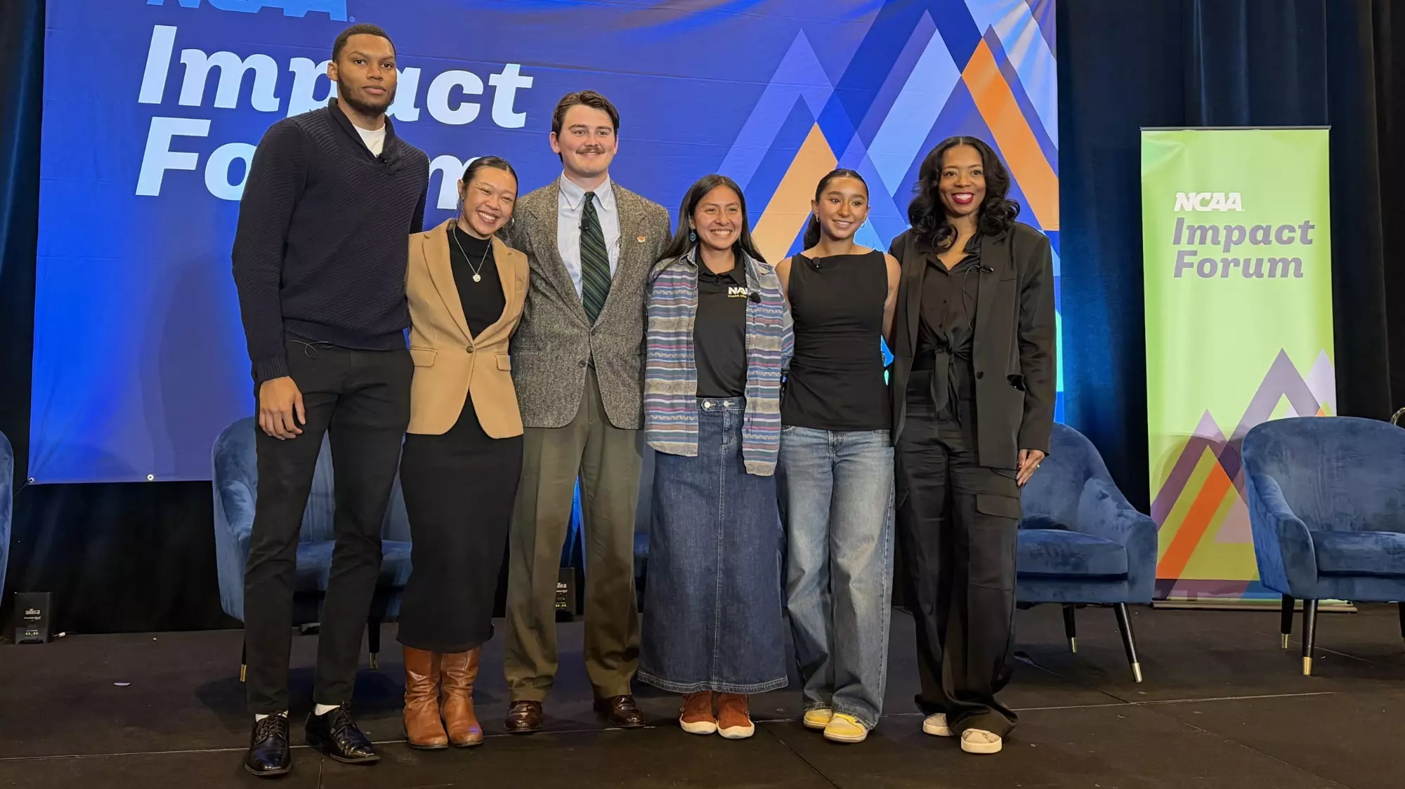 Aidan Hamner stands in a full suit and tie on the stage of an NCAA Impact Forum event, judging by the branding in the back ground of the photo. Hamner stands third from the left in a group of six people. 