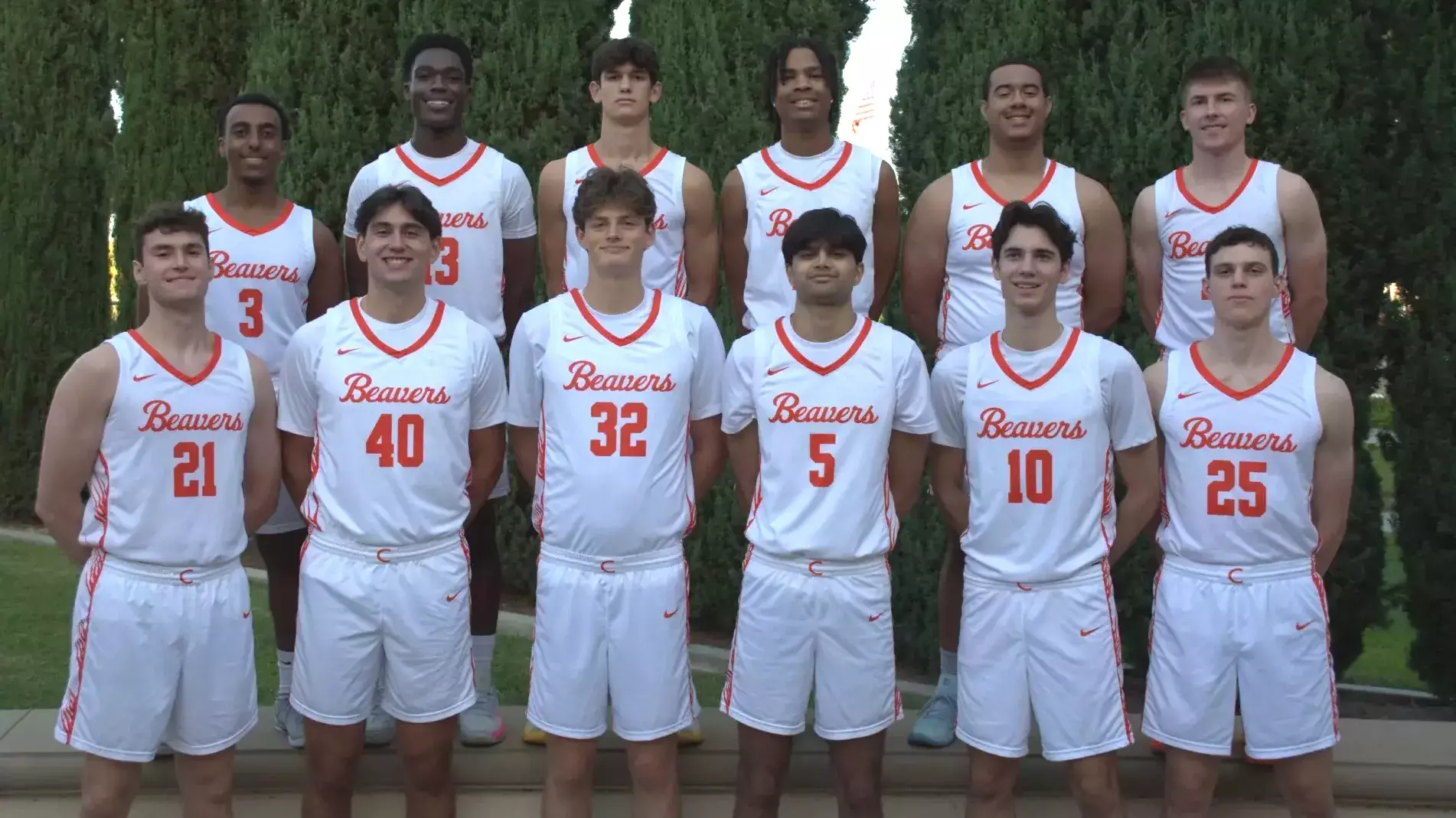 2025-26 Caltech men's basketball stands in front of a row of trees in white uniforms with orange trim. 