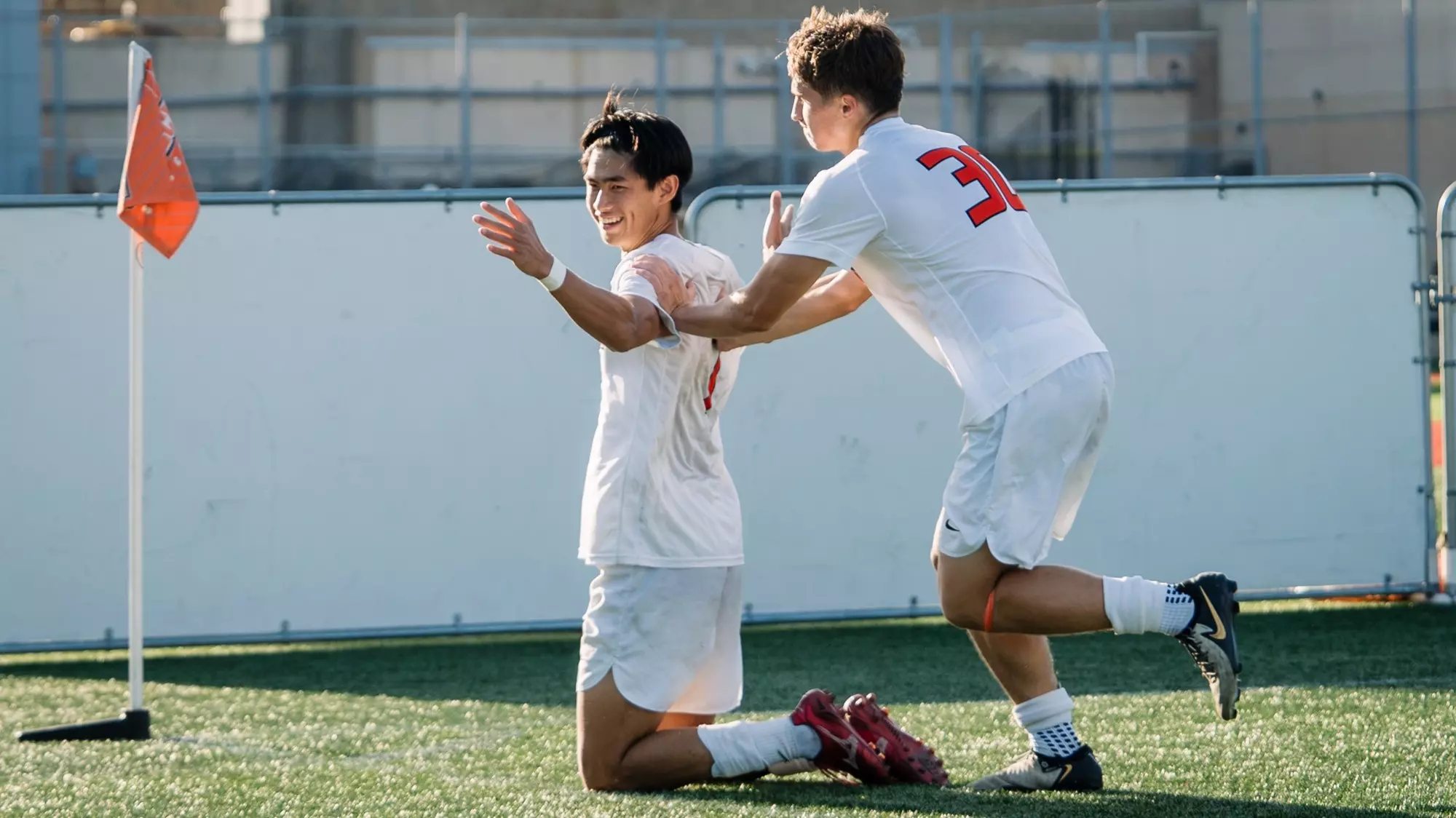 Matthew Luk kneels at the corner flag celebrating a goal as he is congratulated by teammate Sergiu Farca 