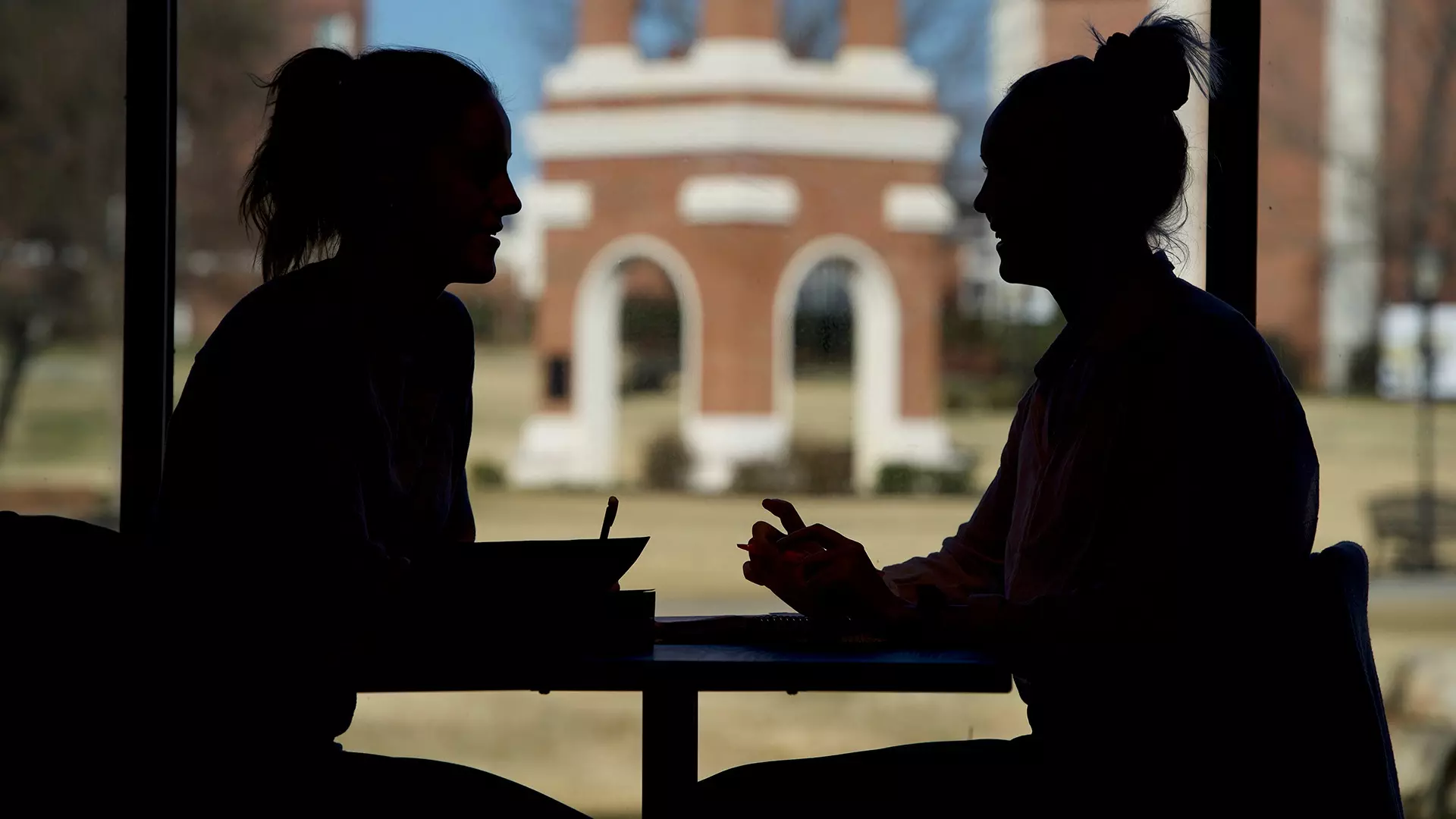 This is a campus life photo of the silhouettes of two students sitting in the library.
