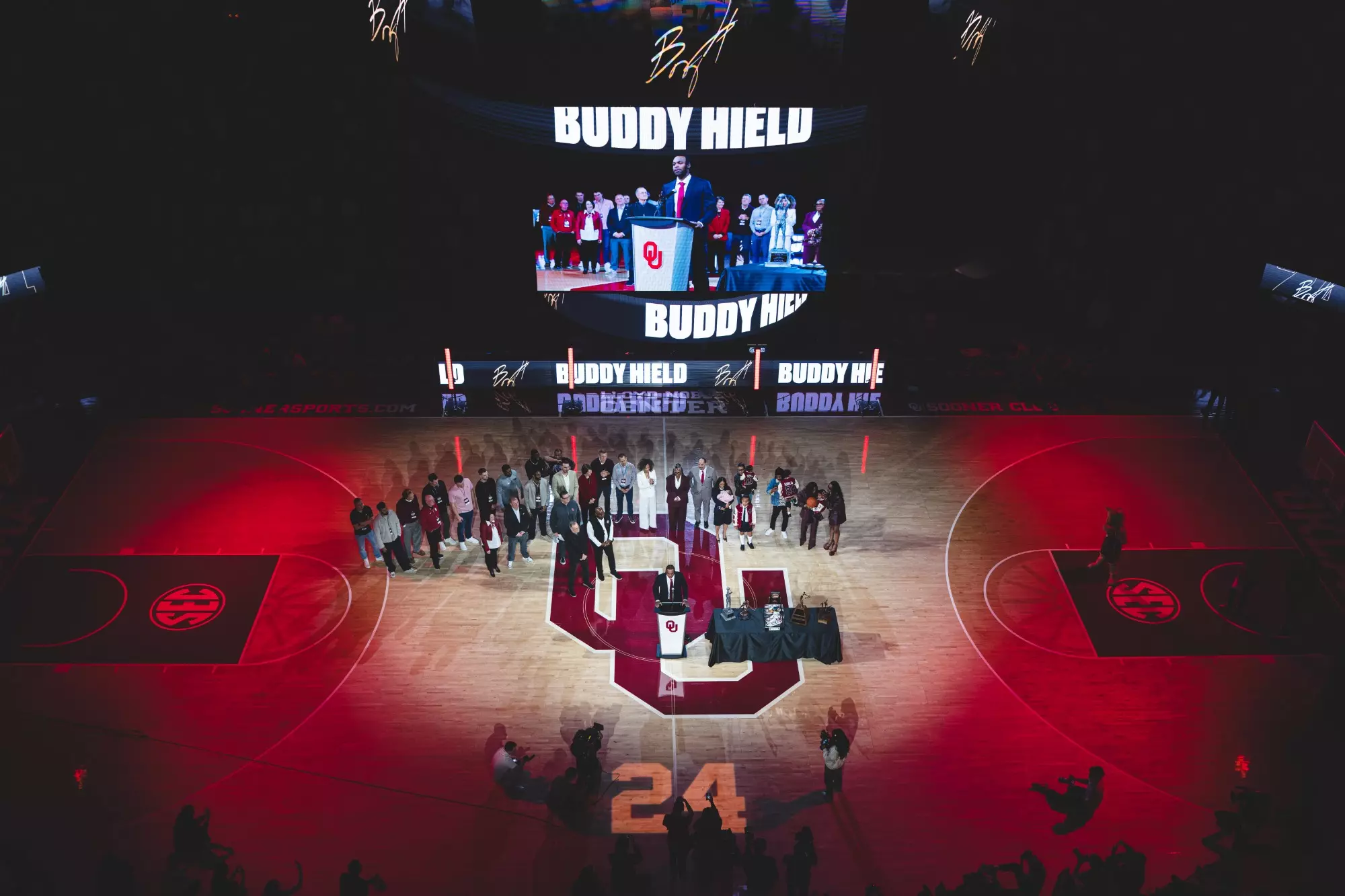 Photo taken from the rafters looking down at group on the court during Buddy Hield jersey ceremony. Court is lit red with number 24 projected on it.