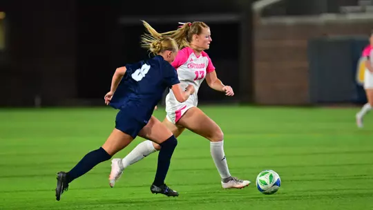 Chattanooga Mocs player in a white uniform dribbles the ball up the field while a Samford defender runs alongside during a women's soccer match.