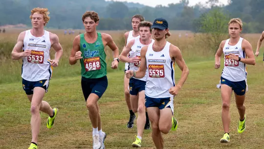 Chattanooga cross country runners compete in a pack during a grassy cross country race, with several Mocs athletes in white uniforms running alongside competitors on an open course.