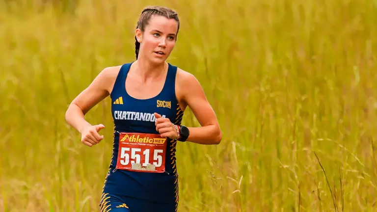 A female runner wearing blue and hair in a French braid is running on a cross country course with tall, brown grass in the background.
