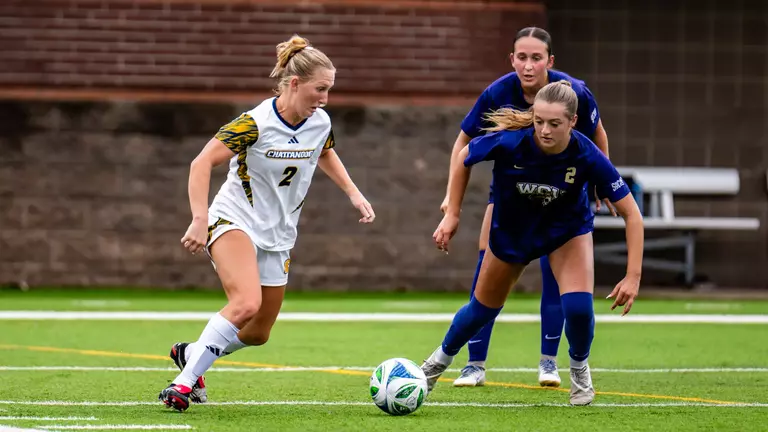 A soccer player in white is taking control of the ball against a defender in dark colors.