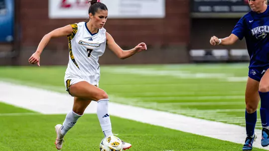 A soccer player in white is controlling the ball while a soccer player in dark is nearby.