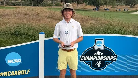 Camden Braidech stands proudly in front of the NCAA Men's Golf Championships signage holding his NCAA Elite Scholar-Athlete Award