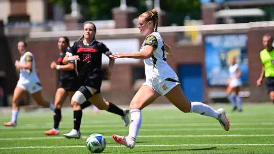A women's soccer player in a white uniform dribbles the ball upfield during a match while two defenders in black uniforms pursue. The play takes place on a sunny afternoon with other players and an official visible in the background.
