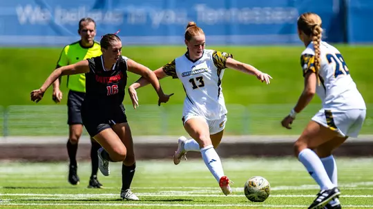 A soccer player wearing white is dribbling the ball while being pursued by a soccer player in black on the pitch.