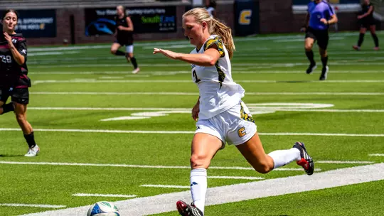 A soccer player in white is running down the field dribbling a ball with her feet.