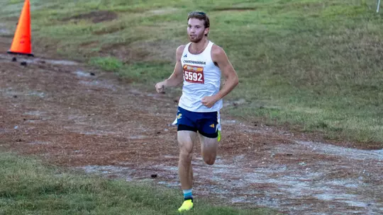 A man is running in a college cross country race on a grassy course.