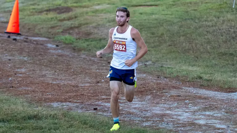 A man is running in a college cross country race on a grassy course.