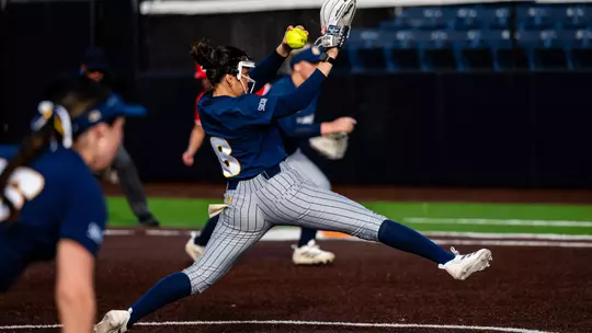A softball pitcher in a blue top and gray pinstripe pants is pitching the ball with her left hand.