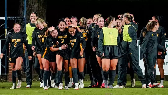 Baylor soccer players in black jerseys with gold lettering smile and embrace on the field at night, gathering around a player wearing No. 10 while teammates and staff in neon yellow bibs and dark warmups celebrate behind them.