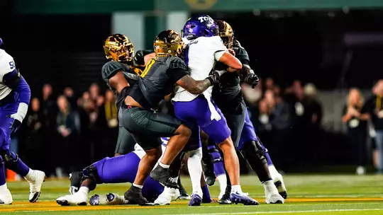 A Baylor defender in a gold helmet and dark gray uniform wraps up a TCU ball carrier in a white jersey and purple helmet during a night game. Another Baylor player closes in from behind as TCU linemen in purple pants are pushed back around them, with a blurred crowd in the stands behind the field.