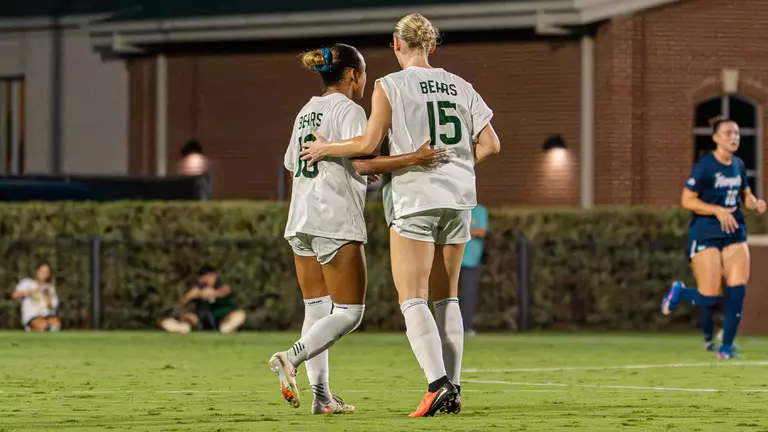 Baylor's Aryanna Jimison (No. 10) and Grace White (No. 15) walk arm in arm across the field during a night match, with the opposing team's colors visible on a nearby player in the background.
