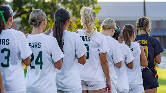 Soccer players in white 'Bears' jerseys stand arm-in-arm in a line facing away from the camera, next to a player in a dark 'Sic 'Em' jersey, before a match on a grass field with trees and a building in the background