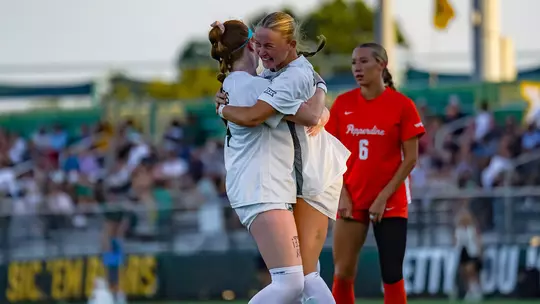 Two Baylor soccer players in white jerseys embrace and celebrate on the field, one facing away from the camera with a red-and-white headband, the other facing the camera smiling. A Pepperdine player in an orange jersey (No. 6) stands nearby in the background, with a crowded stadium behind them.