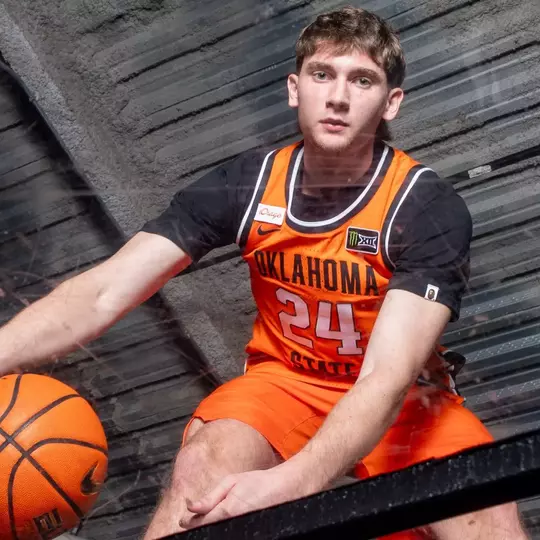 Oklahoma State Cowboy Basketball player Ryan Crotty (#24) poses for a promotional photo beneath a glass backboard, holding a Nike basketball with one hand while looking directly into the camera. He wears the team's orange Oklahoma State jersey with the Big 12 conference logo visible on the chest. The photo is taken from a low angle looking up through the backboard, with orange support structures framing the shot.