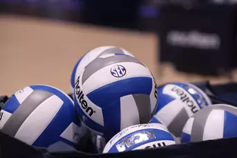 A blue and white volleyball with a conference logo in the center of a basket of volleyballs.