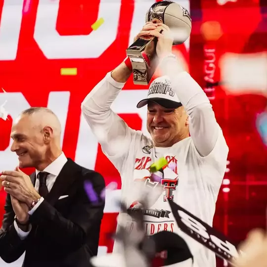 Texas Tech head coach Joey McGuire smiles and raises the Big 12 championship trophy overhead on the podium as Big 12 Commissioner Brett Yormark applauds beside him with confetti falling in front of a red championship backdrop.
