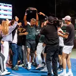 Matt Forbes raises the 2025-26 Men's Tennis Big Ten Tournament trophy while getting swarmed by teammates
