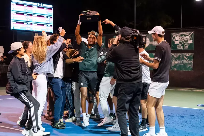 Matt Forbes raises the 2025-26 Men's Tennis Big Ten Tournament trophy while getting swarmed by teammates