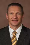 Head Football Coach Jeff Monken Headshot with a black suit and gold tie on.