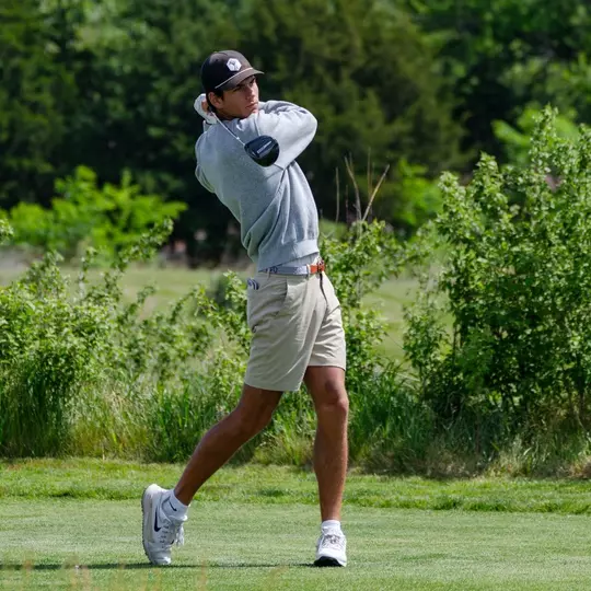 Sam Udovich swings his driver with a background full of green trees.