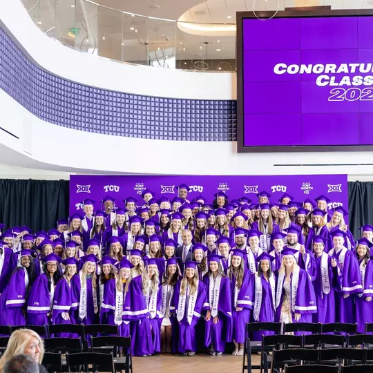 TCU athletics Class of 2026 posing for a photo in their graduation gowns with Athletic Director Mike Buddie
