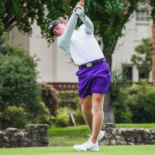 Charlie Wylie swings an iron off the tee in a white shirt and purple shorts