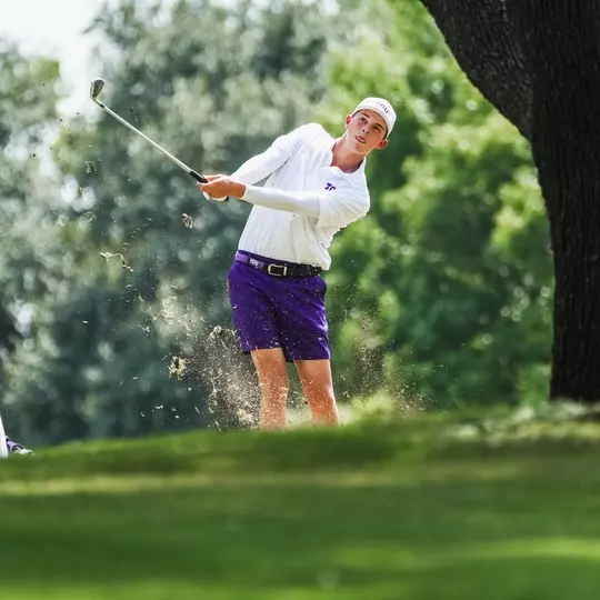 Nathan Miller swings a club under a tree wearing purple shorts and a white shirt