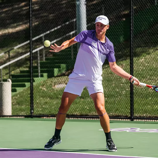 Maximus Dussault coils as he prepares to hit a forehand wearing a white and purple shirt.