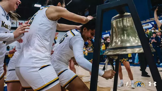 Kent State women's basketball players celebrate a win by ringing the victory bell courtside, teammates cheering and reaching in as one player pulls the rope, with cheerleaders and fans visible in the background.