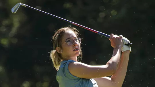 Kent State women's golfer Petra Babicová follows through on a swing during competition, wearing a blue sleeveless top and dark shorts, with trees blurred in the background.
