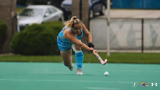 Emily Robertson slapping a pass across the pitch to a teammate as water flies off her stick from the grass