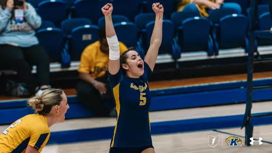 Kent State volleyball player #5 in a navy and gold jersey raises both fists overhead and yells in celebration, with a white wrap on her forearm. A teammate in a gold jersey leans in at the lower left, also cheering, while fans sit in the blue bleachers behind them.