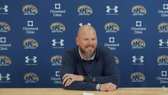 A bald man with a beard smiling while seated at a wooden desk in front of a blue Kent State press backdrop featuring repeating logos for Kent State, Under Armour, and Cleveland Clinic.