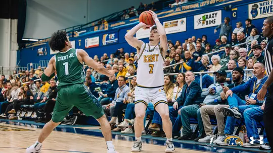 Kent State player #7, in a white home jersey with gold and navy trim, rises into a three-point shooting motion with the ball overhead while Emery Jr. (#1) of the opposing team, in a green jersey, defends. Fans fill the stands behind them.