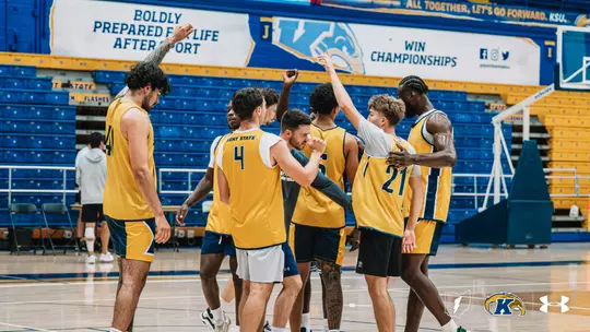 Kent State men's basketball players in gold practice jerseys gather in a tight huddle at center court, with several raising an arm into the air, in an otherwise empty arena with blue seating.