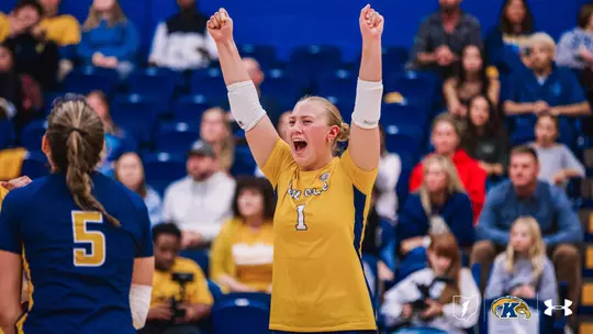 Kent State volleyball player in a gold No. 1 jersey raises both fists above her head and yells in celebration, while a teammate in a navy No. 5 jersey stands in the foreground with her back to the camera. Fans fill the blue bleachers behind them.