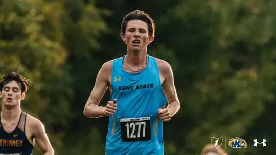 Kent State men's cross country runner in a light blue singlet with bib number 1277 runs toward the camera with his fists clenched and a silver chain around his neck, while a competitor in a navy and gold uniform trails behind him. Green trees fill the background.