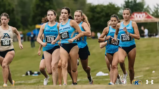 A pack of Kent State women's cross country runners in light blue singlets, including bib numbers 1376, 1377 and 1378, runs across a grassy hillside beside a runner in a cream Akron uniform with bib number 1367. Event tents are visible in the background.